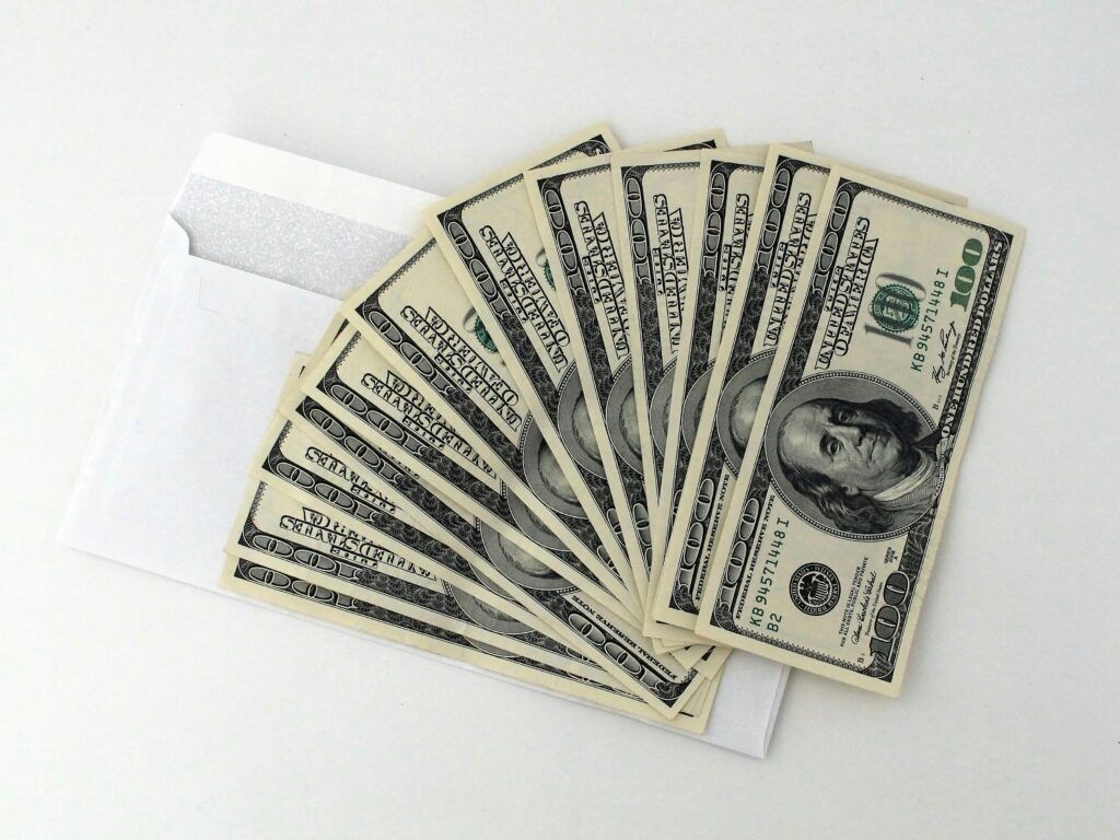 Fan of US $100 bills partially out of a white envelope on a white background. Comparing payment methods and payout frequency for affiliate programs.