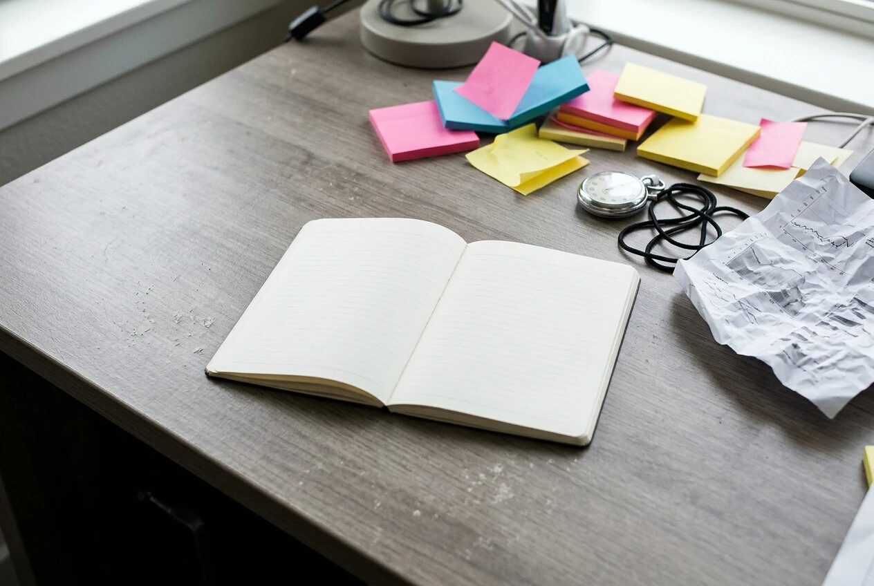 A cluttered desk corner with a stopwatch, a tangle of unused colourful sticky notes and a crumpled printed graph pushed to the edge, while a single clean notebook sits open in the centre of the desk, bright daylight, muted greys with a few bright accents.
